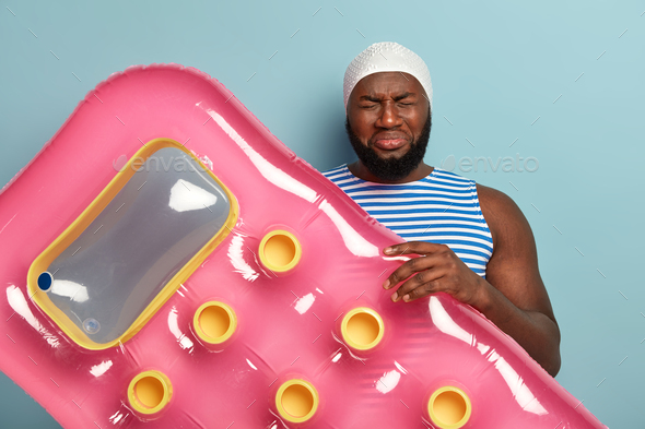 Unhappy man with dark skin, holds floating mattress, wears special swimhat, being in low spirit beca