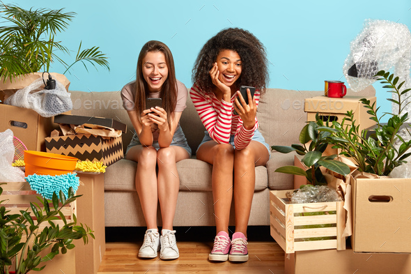 Mixed race women rest on sofa with unpacked boxes around after moving into new house, rent flat toge