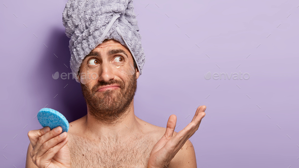 Studio shot of puzzled doubtful man uses cotton disk to apply lotion on face, removes wrinkles with