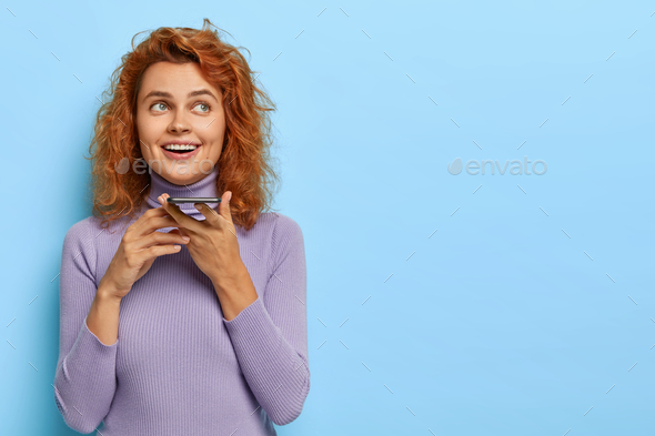 Pleased ginger girl makes voice call, holds smartphone near mouth, glad to have good talk with frien