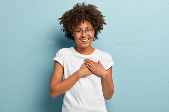Indoor shot of happy dark skinned lady swears or promises something, holds hands on chest, tells tru