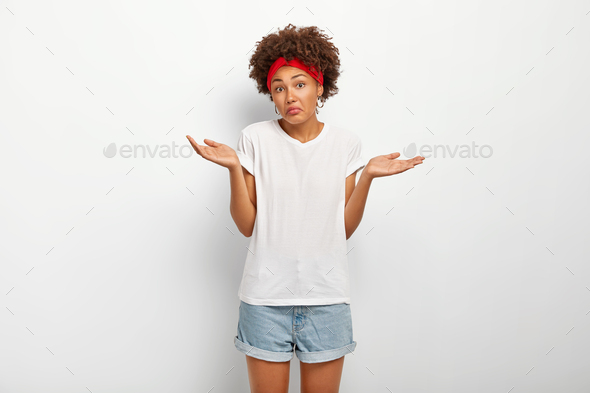 Uncertain clueless female with Afro hairstyle, spreads hands with doubt, cannot make decision, faces