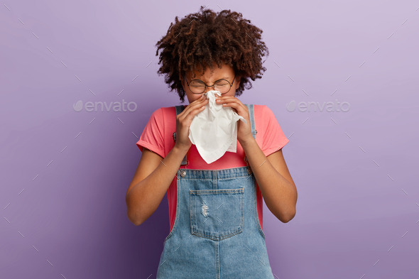 Curly young female feels unwell, blows nose in white tissue, suffers from running nose, cold symptom