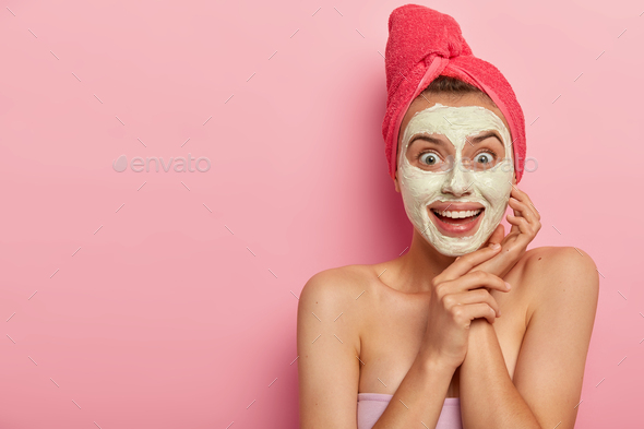 Happy joyful young woman applies cream mask wonderful for any skin type, wears red towel on head, no