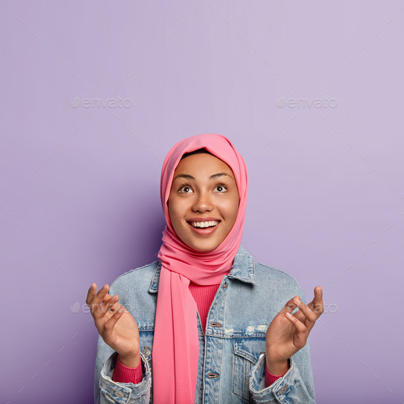 Religious cheerful woman has Islamic views, raises palms, prays and looks hopefully upwards, wrapped