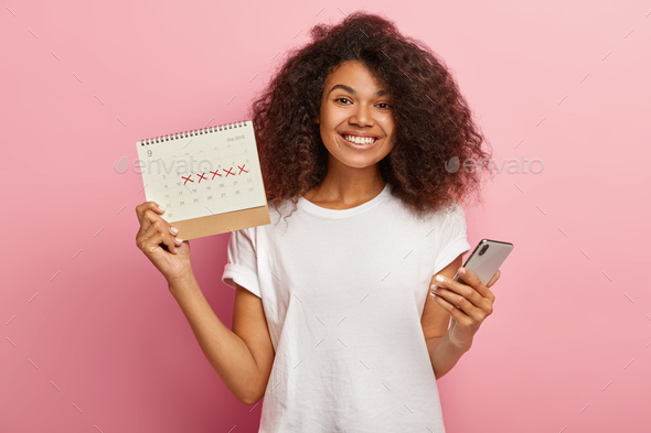 Happy curly woman holds periods calendar, checks menstruation days on mobile phone application, care