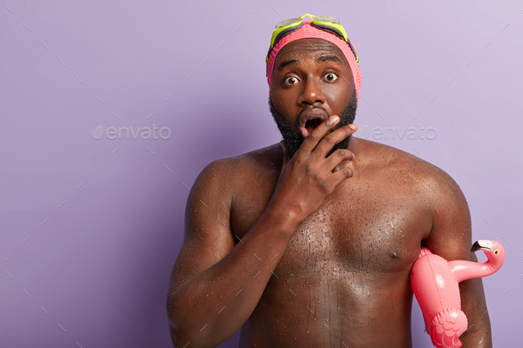 Waist up shot of embarrassed dark skinned guy opens mouth widely, has muscular body, swims in swimmi