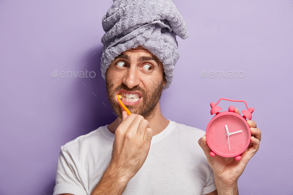 Handsome man brushes teeth, whitens with toothpaste, holds alarm clock in hand, awoke late in mornin