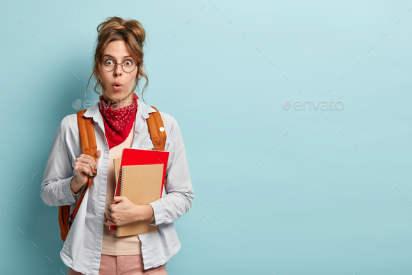 Impressed stupefied pupil attends language courses, holds notepads, wears glasses, red bandana and s
