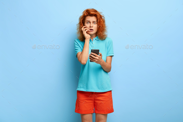 Puzzled displeased redhaired woman shocked not to receive email confirmation, holds modern cell phon