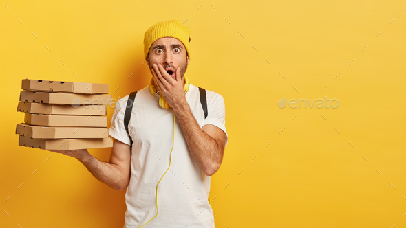 Portrait of young shocked male delivery worker holds stack of pizza boxes, dressed casually, covers