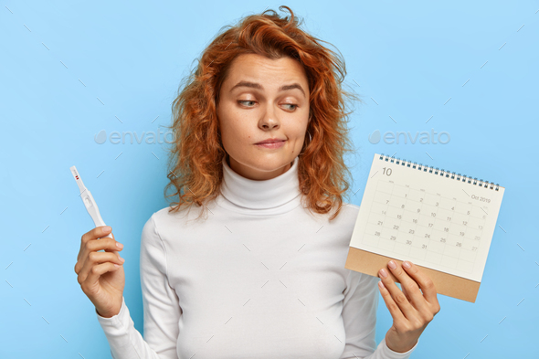 Image of attractive woman holds pregnancy test and periods calendar, checks menstruation days, finds