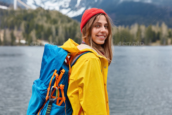 Hiking woman stops near lake in mountains, carries backback, holds thermos  of hot beverage, explores Stock Photo by wayhomestudioo