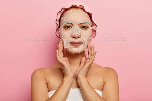 Serious japanese female puts nourishing mask on face, applies moisturizer sheet product on skin, wea