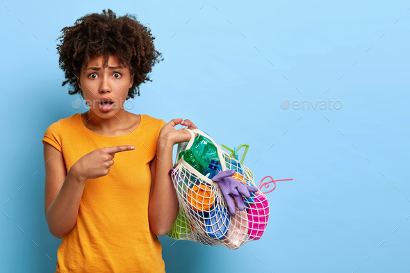 Puzzled worried woman volunteer picks up plastic waste, points at bag with litter, wears orange t sh