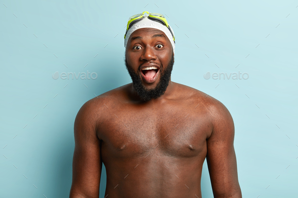Dark skinned handsome guy with naked body, prepares for swim, wears special hat and goggles, keeps m
