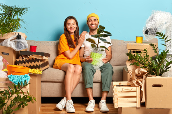 Delighted woman and man sit on sofa with household objects, buy luxury house to start new family lif