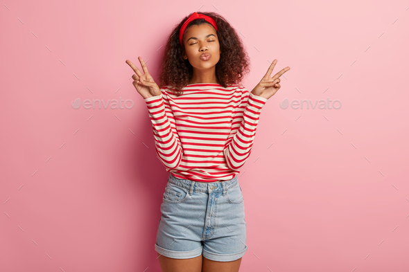 Peaceful pretty beautiful woman with Afro hairstyle, keeps lips folded, makes peace gesture with bot