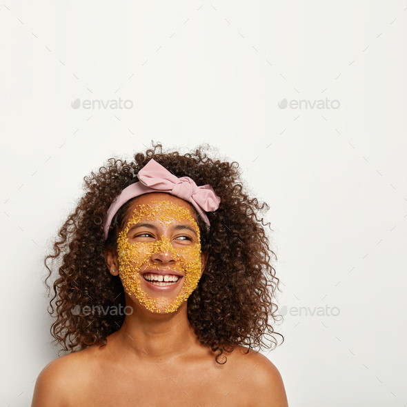 Positive overjoyed woman laughs happily, has beauty procedure, applies yellow granule scrub for anti
