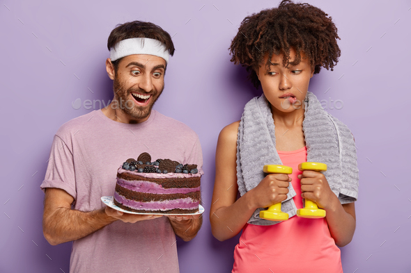 Unhappy Afro female temptates her will power, bites lips as looks at tasty baked cake in mans hands,