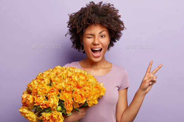 Funny young woman blinks eyes, makes peace sign with hand, expresses happiness, holds orange tulips,