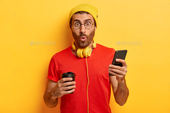 Surprised unshaven guy listens music in headphones, sends text messages on cell phone, types answer,