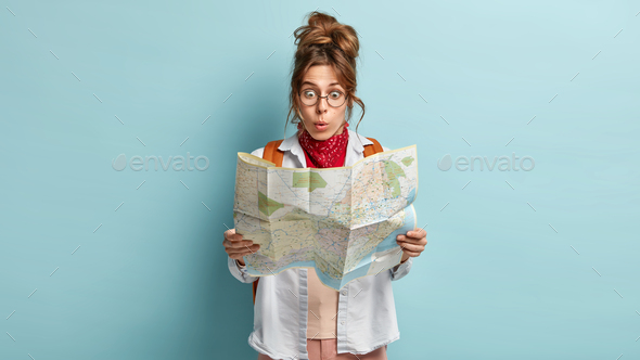 Photo of shocked traveler lost way, tries to find destination, stares at paper map, stares through r