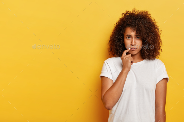 Sad sulking Afro American millennial woman, keeps index finger near corn of lips, insulted by someon
