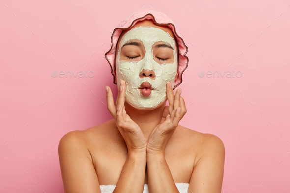 Close up image of pleased woman applies homemade facial mask for dry skin, makes fish mouth, has spa