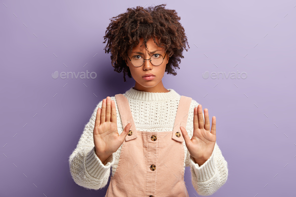 Dissatisfied young woman with dark skin, curly hair, makes stop gesture, has angry facial expression