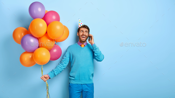 Festive glad man has pleasant friendly conversation via cell phone, carries bunch of balloons, invit