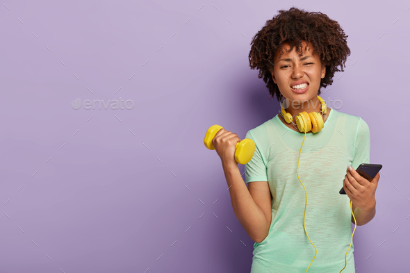 Dissatisfied tired woman dressed in sports clothing, raises hands with kettlebell, smirks face, hold
