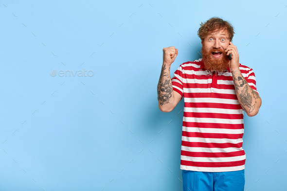 Overjoyed happy ginger male winner celebrates getting new job opportunity, clenches fists while talk