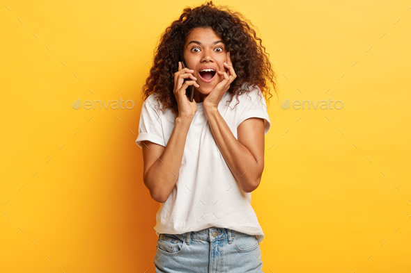 Sociable friendly impressed curly African American teenage girl holds breath from awesome news, call