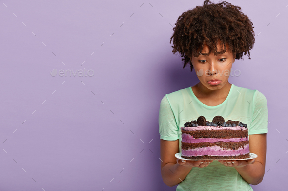 Photo of dissatisfied Afro American woman holds plate of blueberry sweet cake, purses lower lip, has