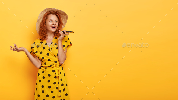 Horizontal studio shot of cheerful young woman makes voice call, holds up to date cell phone, enjoys