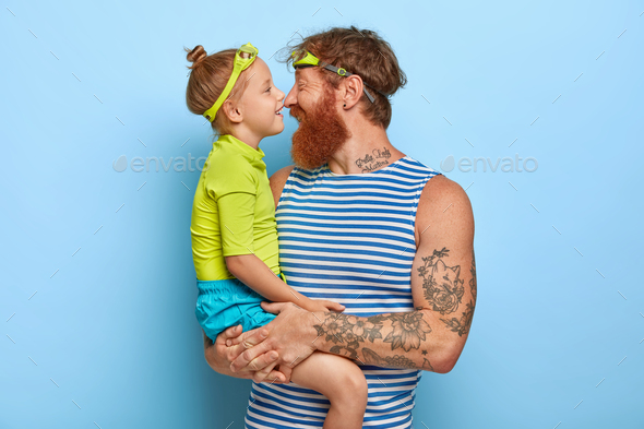 Photo of bearded redhead young man carries little daughter, touch noses and have happy expressions,