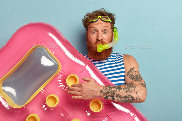 Studio shot of surprised bearded red man wears snorkel mask, holds pink inflated mattress, wears sai