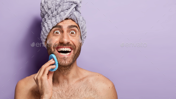 Glad handsome man uses cosmetic sponge and under eyes pads after taking shower, cleans face, smiles