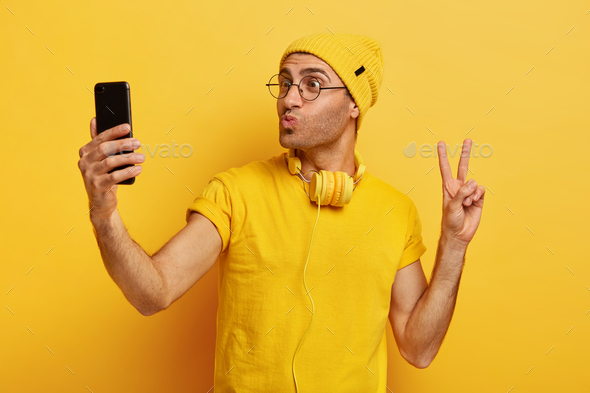 Handsome stylish hipster takes selfie, makes peace gesture, keeps lips folded, holds smart phone, we