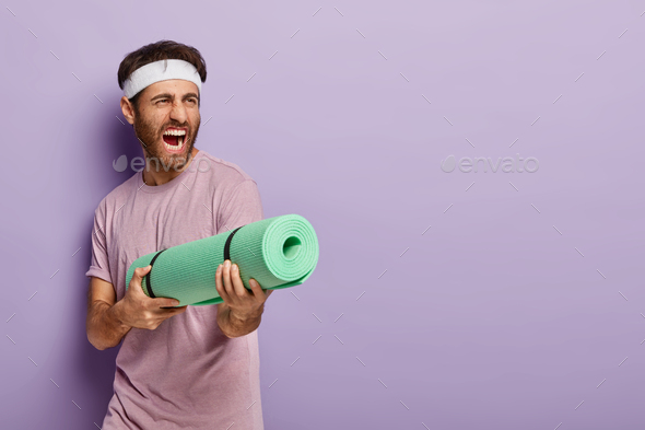 Emotive handsome man pretends shooting in someone with fitness mat, screams loudly, has fun after yo
