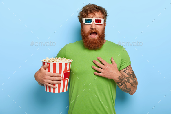 Emotional red head man looks with great fright, watches horror film alone in cinema, has bated breat