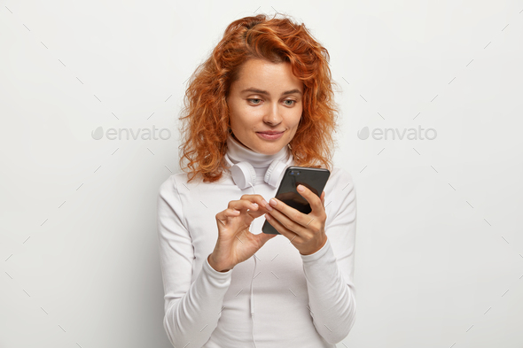 Attentive woman focused at smartphone device, makes song playlist, downloads app, has modern headpho