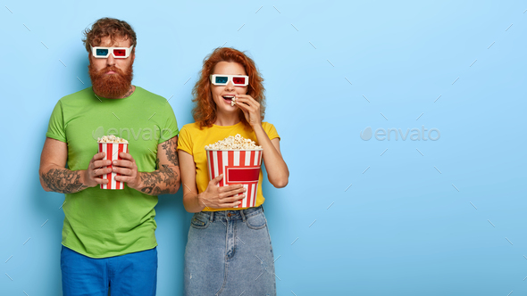 Happy ginger woman and sad man wears cinema glasses, watch film premier, eat delicious popcorn toget