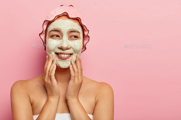 Glad young woman with clay facial mask, gets spa procedures in beauty salon, pampers face, stands wr