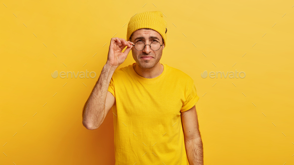 Discontent male model looks thoroughly through round glasses, has bad eyesight, tries to notice some