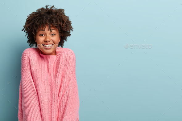 Positive dark haired female gazes happily with tender smile, dressed in oversized rosy sweater, plea