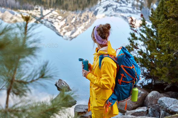Hiking woman stops near lake in mountains, carries backback, holds thermos  of hot beverage, explores Stock Photo by wayhomestudioo