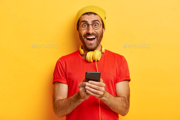 Indoor shot of optimistic Caucasian man downloads new app for listening music on mobile phone, smile - Stock Photo - Images