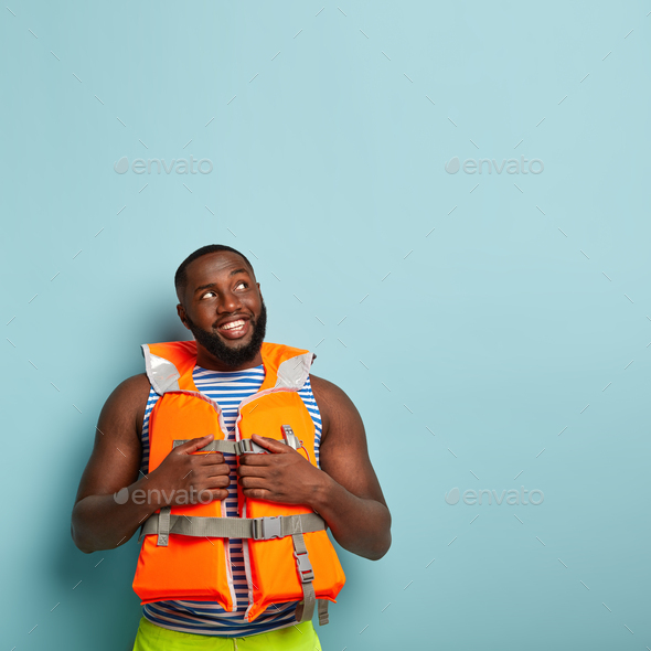 Positive African American man happy to have nice entertaining cruise, wears orange lifejacket, has s